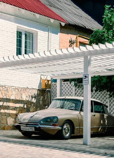 Carport à claire-voie en bois blanc adossé à une maison en pierre, véhicule ancien garé dessous
