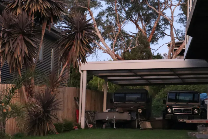 Deux véhicules 4x4 garés sous un carport aluminium double à claire-voie, au crépuscule dans un jardin arboré