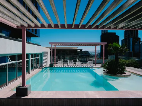 Pergola bioclimatique autoportée rose au bord d’une piscine sur un toit-terrasse, vue sur la ville